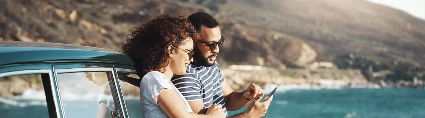 Woman and man leaving against a car smiling at phone next to the ocean